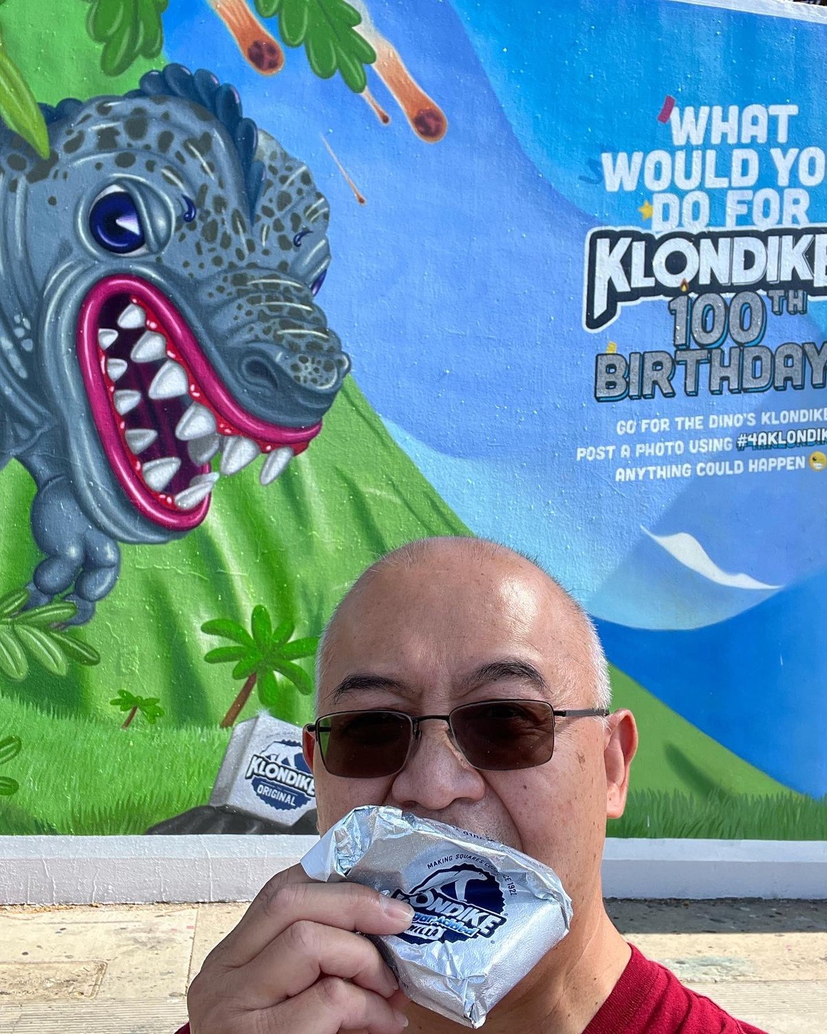 A visitor biting into a Klondike bar in front of the dinosaur and the 100th birthday headline