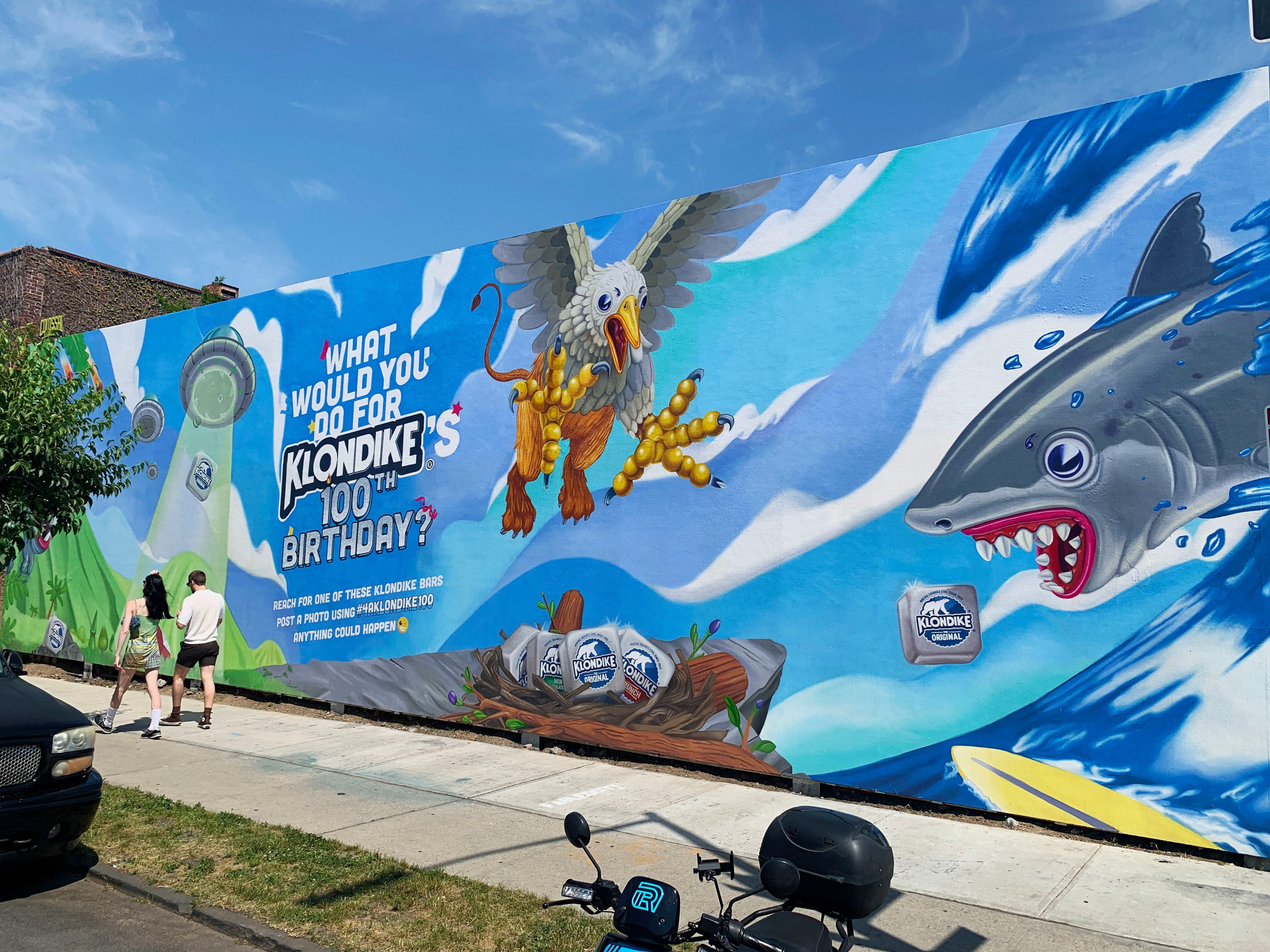 The finished Klondike mural along a Brooklyn sidewalk, two people walking past the griffin and the shark
